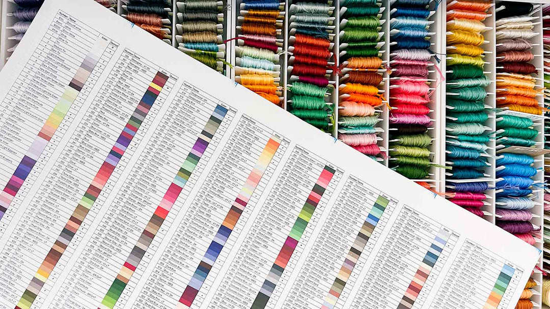 A printed thread inventory spreadsheet with color swatches and numbers lays diagonally over a drawer filled with rows of neatly organized, colorful embroidery floss bobbins.