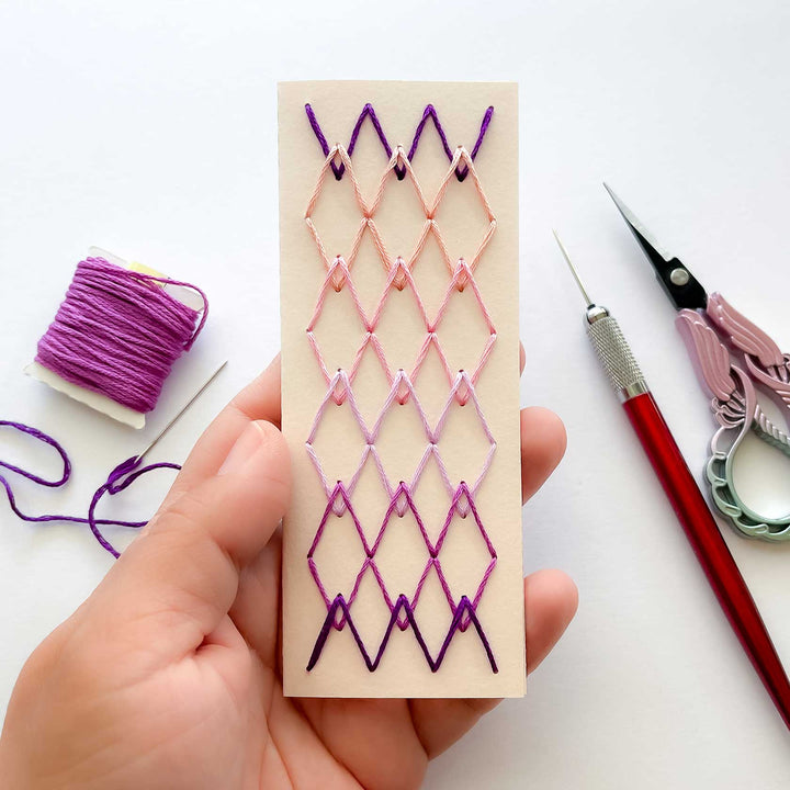 Handmade cross-stitch bookmark with purple and pink thread, surrounded by crafting tools on a white background.