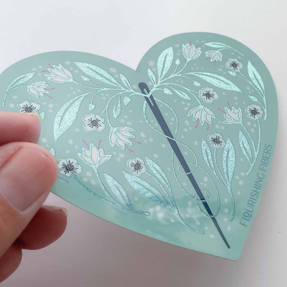 Heart-shaped sticker with floral design and needle held by a hand