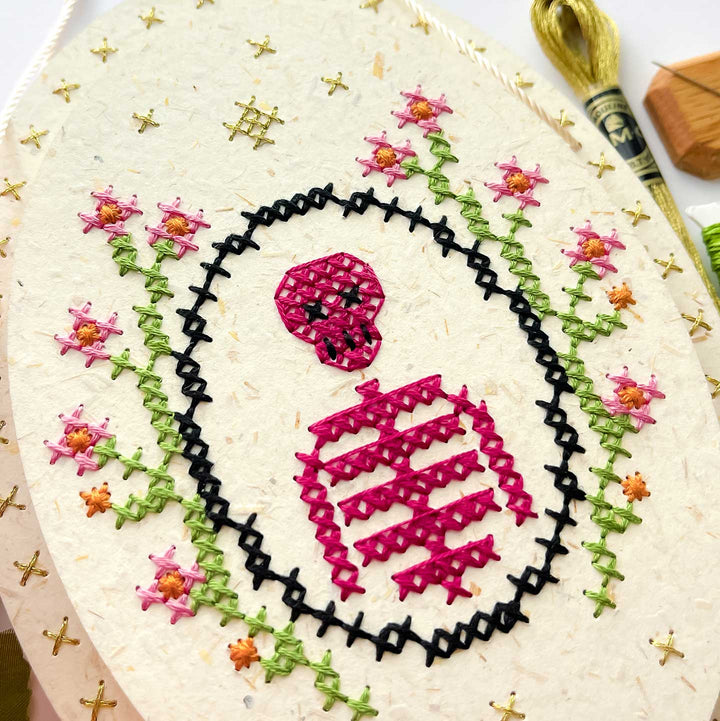 A close-up of a hand-embroidered oval textile featuring a pink skull and crossbones design inside a black stitched border, surrounded by pink flowers with green stems on a cream-colored fabric background.