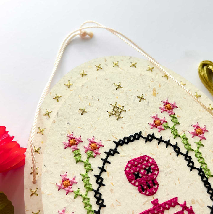 A close-up of a handmade paper hanging ornament featuring a sugar skull design with pink flowers, green stems, black stitched details, and gold star accents, suspended by a cream cord.