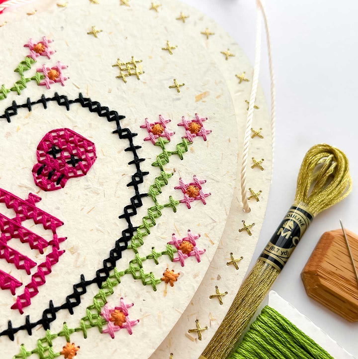 A close-up of a circular embroidery canvas with a partially completed floral design in black, green, pink, and gold thread, alongside a spool of gold thread, a wooden needle block, and a needle on a white background.