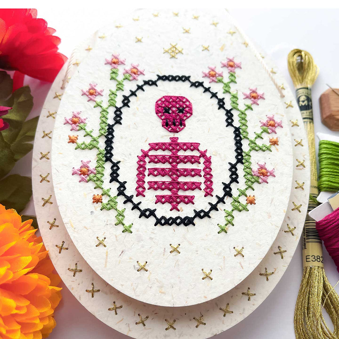 A decorative embroidery featuring a pink stitched skeleton and skull motif inside a black oval, surrounded by small pink and green flowers. The piece is displayed on stacked white paper pads with gold stitched borders, alongside embroidery floss threads and flowers in the background.