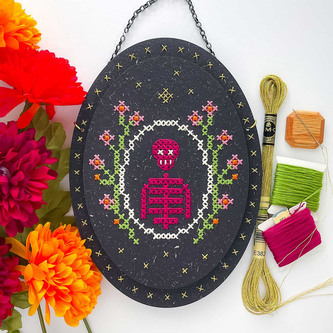 An oval black cross-stitch sampler featuring a pink skeleton design with green and orange floral details, styled with colorful flowers and embroidery threads on a white background.