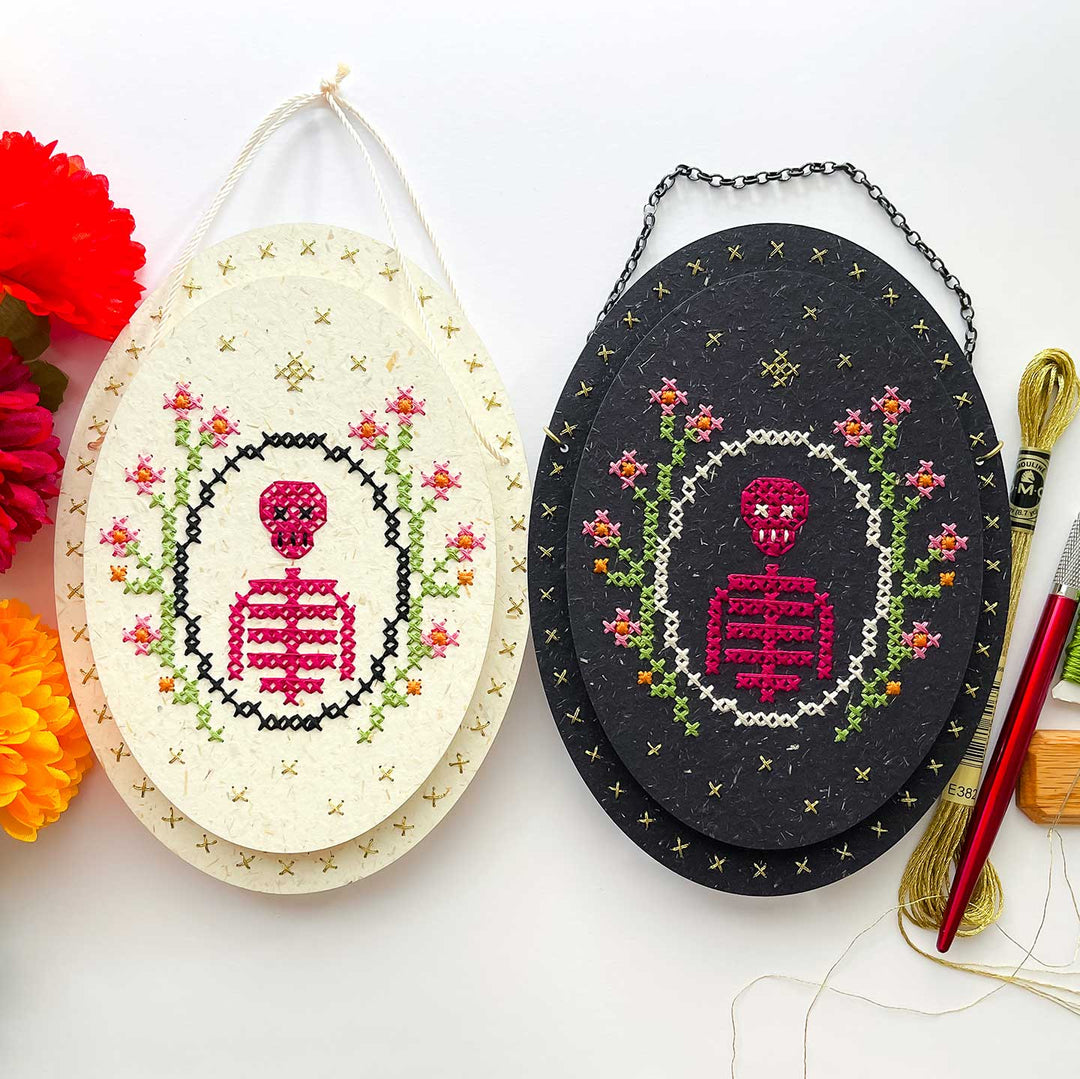 Two oval cross-stitch needlework kits displayed side by side on a white background. The left kit has an off-white fabric with a pink skeleton and floral design, and the right kit has a black fabric with the same design.