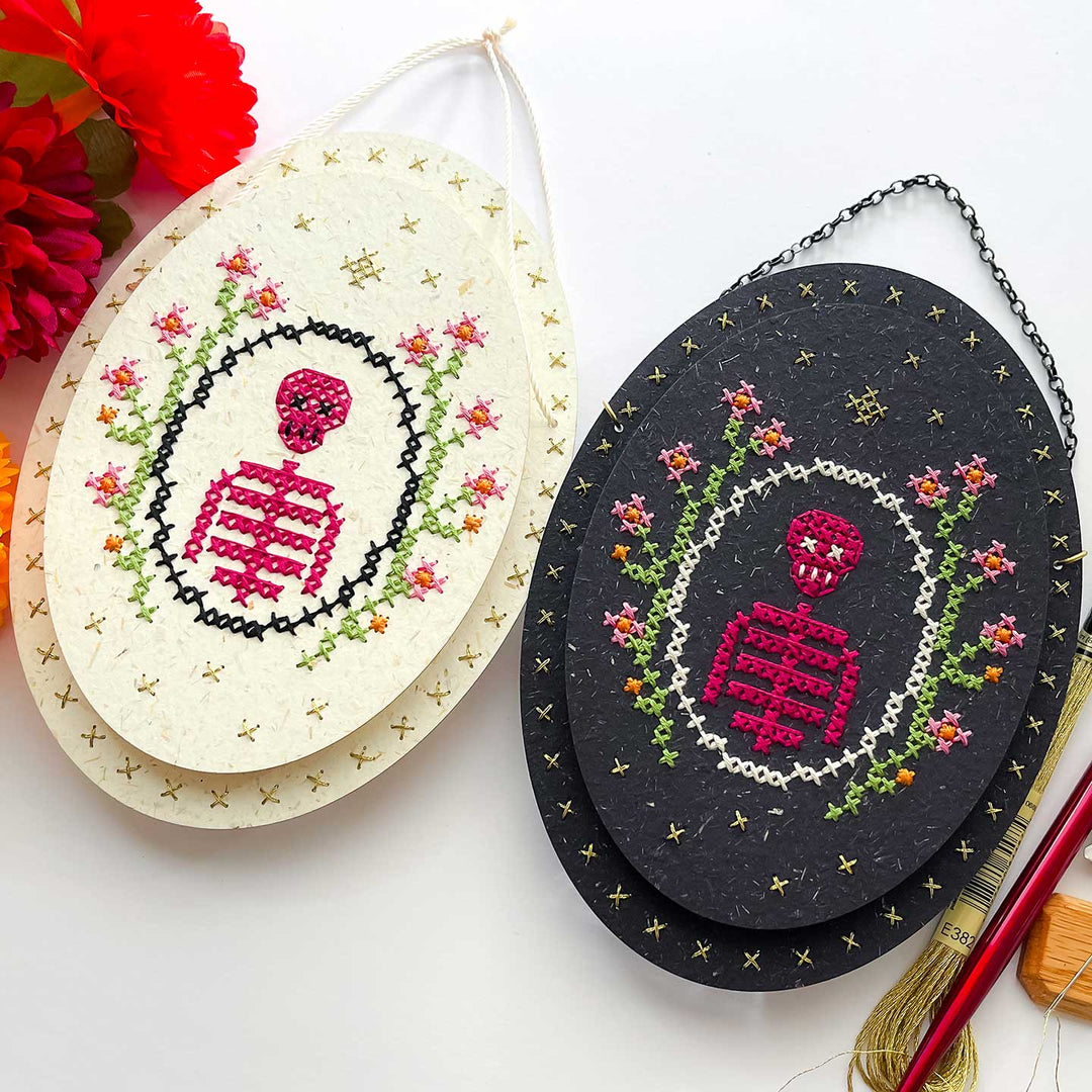 Two oval hanging cross-stitch art pieces displayed on a white background. One has an off-white background with gold star details and the other has a black background with a chain.