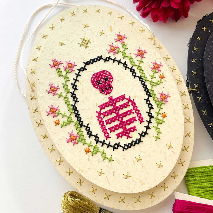 An oval handmade paper cross-stitch sampler with a traditional character design in pink and black, surrounded by embroidered flowers in green and pink, displayed on a white background with other craft supplies partially visible.
