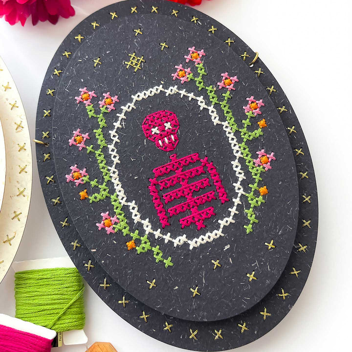 A handmade oval cross-stitch piece on dark charcoal paper, featuring a pink skull and crossbones, green floral vines, and a white border, styled on a white surface with embroidery floss.