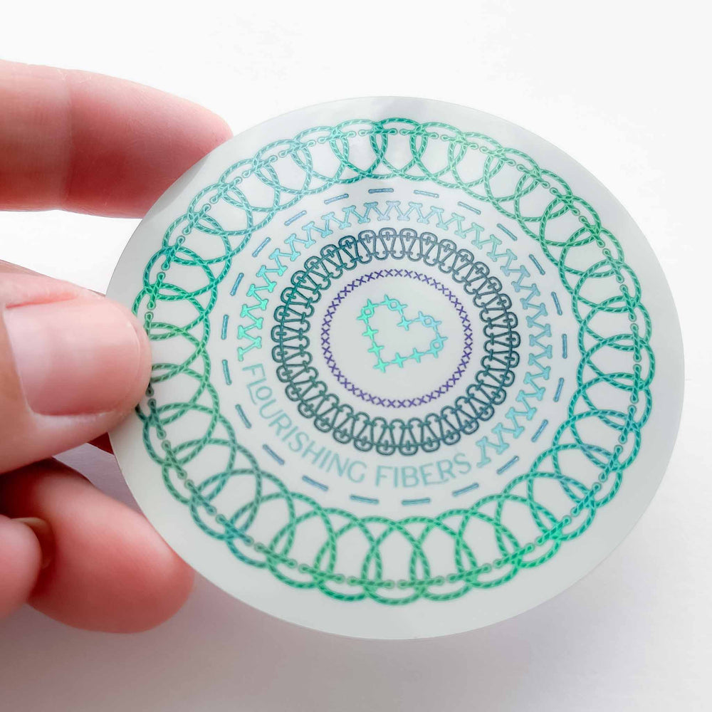 A person's hand holds a circular holographic sticker against a white background. The sticker features concentric rings of various embroidery stitch styles in mint and teal, with a cross-stitch heart at the center and the words "Flourishing Fibers" curved along the bottom.