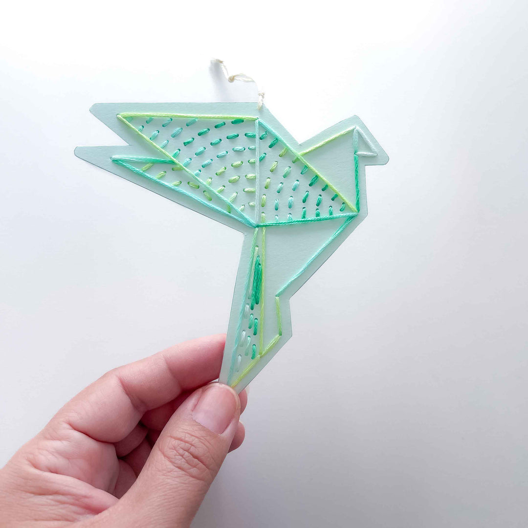 Hand holding a geometric, teal-colored paper embroidery ornament in the shape of a bird against a white background