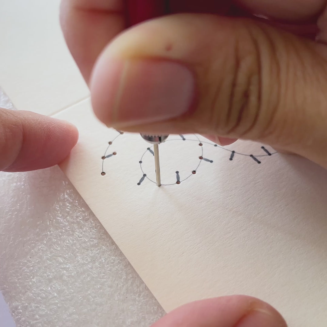 Using awl to pierce holes on a paper card. Embroidery of a line in yellow thread.