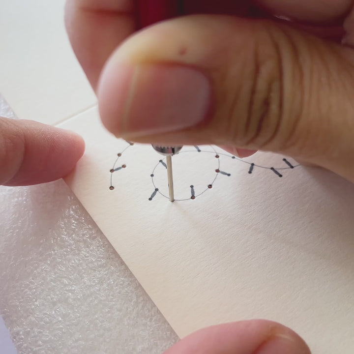 Using awl to pierce holes on a paper card. Embroidery of a line in yellow thread.
