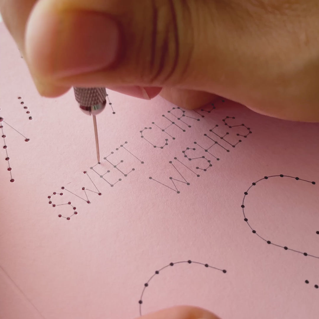 Work-in-progress video demonstrating the paper embroidery process, showing an awl piercing template holes and hand stitching the lettering on a candy cane Christmas card.