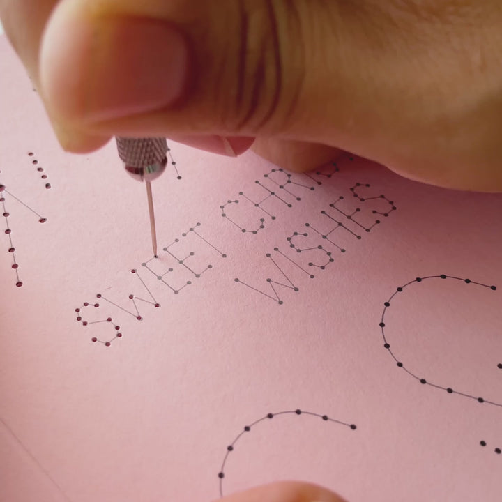 Work-in-progress video demonstrating the paper embroidery process, showing an awl piercing template holes and hand stitching the lettering on a candy cane Christmas card.